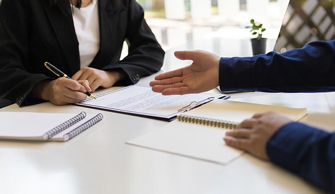 A person in a suit signing a document on a clipboard, sitting across from another person at a desk