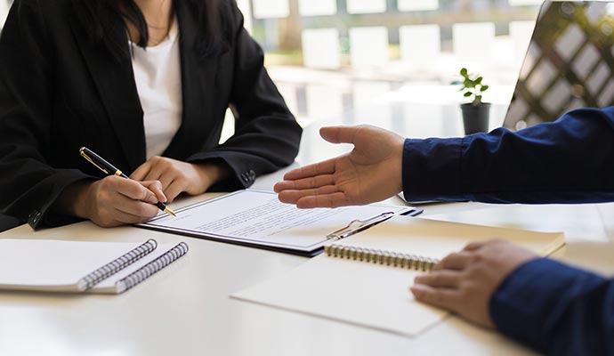 A person in a suit signing a document on a clipboard, sitting across from another person at a desk